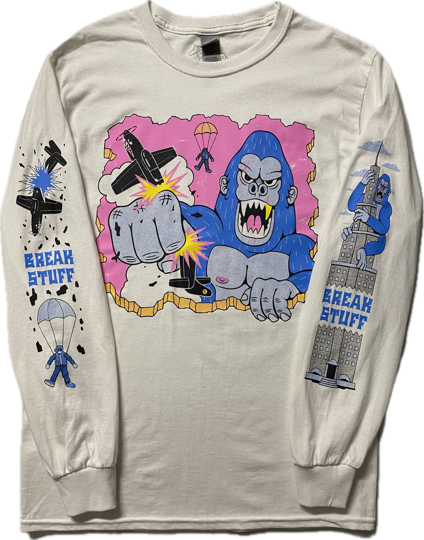 A white long sleeve t-shirt with a cartoon blue gorilla punching a black plane. Behind the gorilla is the ejected pilot. On the right sleeve it reads Break Stuff in blue font with a black broken plane above it and a parachuting pilot below it. On the left sleeve is a blue gorilla climbing a high rise building and it reads Break Stuff too.