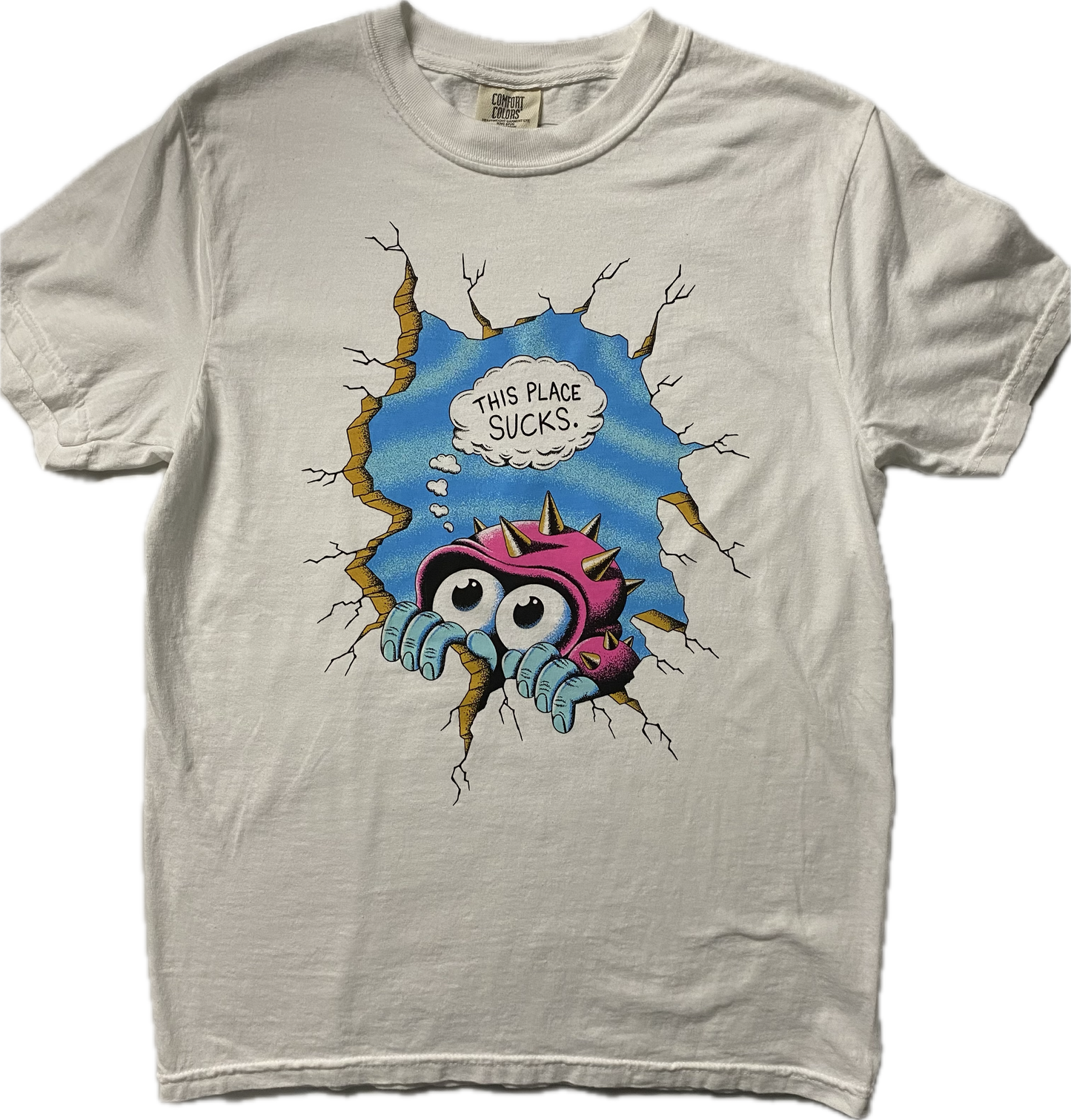 A white short sleeve t-shirt with a cartoon figure peeking over a crack in a cracked wall. It has big googly eyes and is wearing a pink hat with gold spikes, behind it is blue sky. Above the figure it reads This Place Sucks in black font in a white cloud.