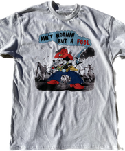 Load image into Gallery viewer, A white short sleeve t-shirt with a cartoon cowboy mouse printed on the front. The mouse has a large red cowboy hat, blue chaps and red boots. It is also wearing white gloves with a red star. It's left had has a pistol pointing upwards. The background has 4 white horses in stampede. Above the mouse it reads Ain't Nothin But a Fool in puffy black and red letters.