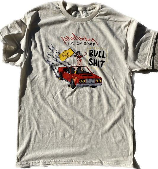 A short sleeve natural colored t-shirt with a chest print of a cartoon girl in a red bikini standing on a red convertible that is driving itself. The car has gold flames above the front tires and smoke coming off the back wheels. The girl is holding a yellow flag with a smiley face on it. Above her reads I'm On Some Bull Shit with red outline flames on top.