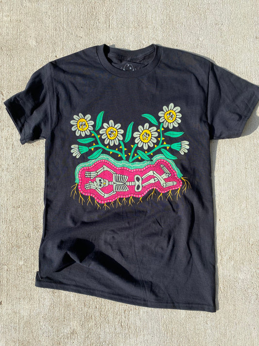 Black short sleeve t-shirt with 6 cartoon flowers with white petals and smiling faces. Below the flowers is a skeleton laying down in pink dirt and roots growing out of the bottom.