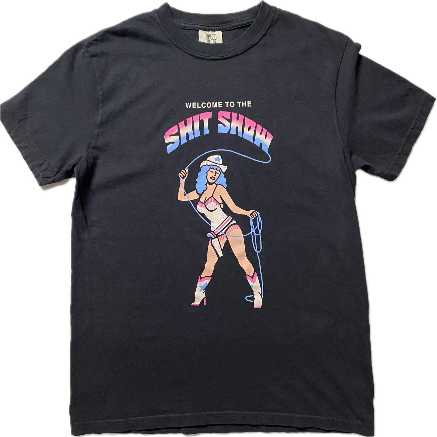 A black short sleeve t-shirt that reads Welcome to the Shit Show. The last 2 words are in a large font in rainbow colors of light blue, white and pink. Below the words is a cowgirl with blue hair in a white bathing suit and white boots. She is holding a blue lasso and have a holster with a pistol.