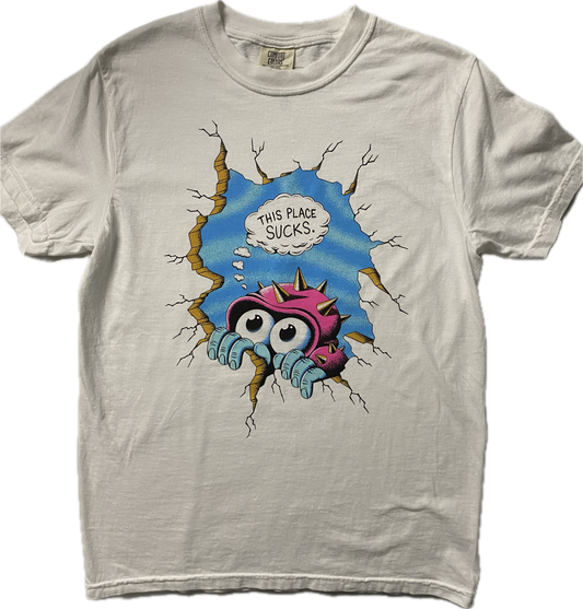 A white short sleeve t-shirt with a cartoon figure peeking over a crack in a cracked wall.  It has big googly eyes and is wearing a pink hat with gold spikes, behind it is blue sky.  Above the figure it reads This Place Sucks in black font in a white cloud.