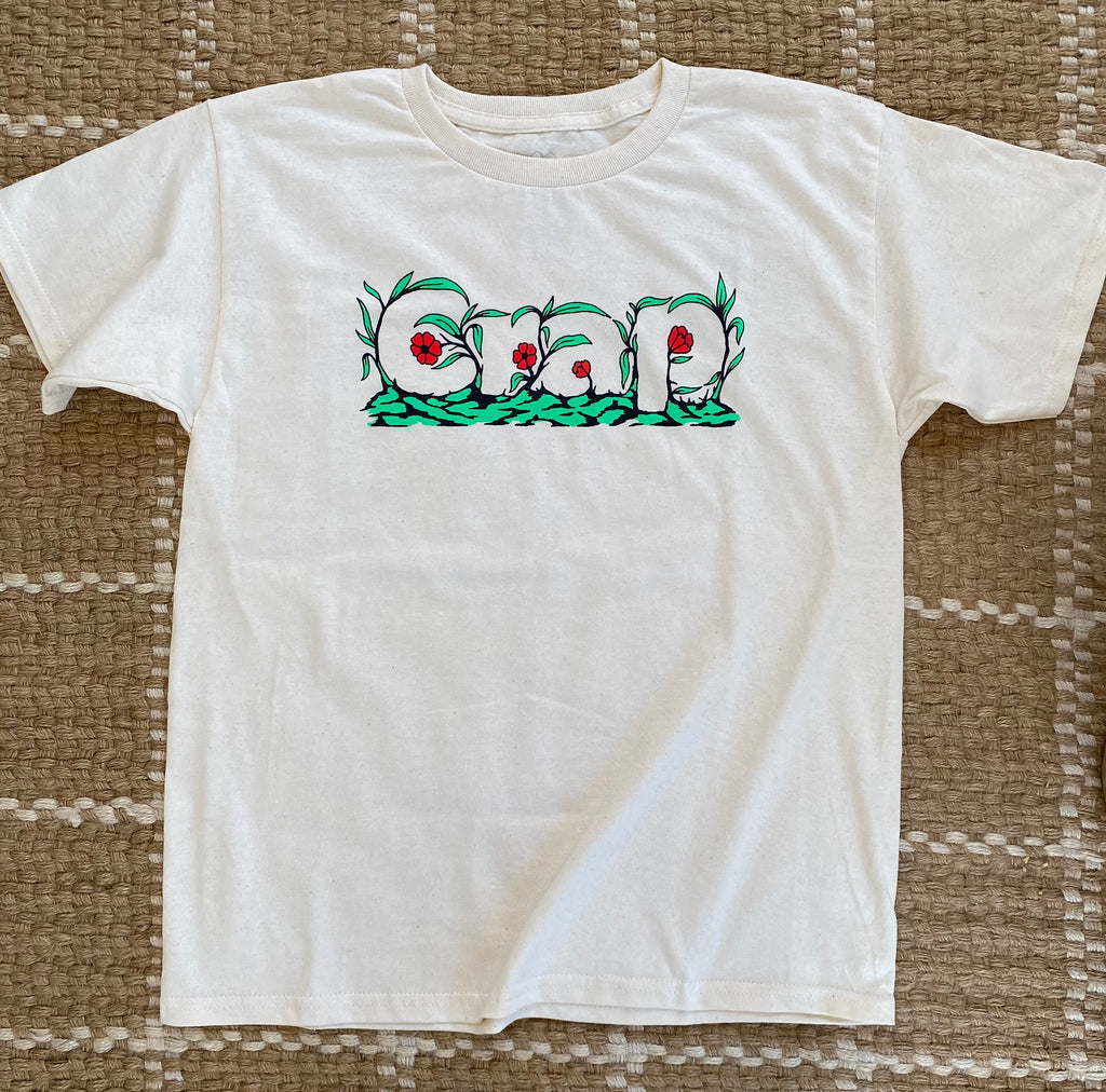 A cream colored short sleeve tee with the word "Crap" printed on the chest area that is camouflaged in green leaves and red flowers.  Tan jute rug as background.