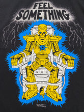 Cargar imagen en el visor de la galería, A close up of the artwork on a black short sleeve tee that reads Feel something at the top with silver color and lighting around it. Below the words is a cartoon skull robot in gold that is strapped in an electric chair. Between it&#39;s feet is a gold mouse coming out of the bottom of the chair. Crocodile Jackson signature at the bottom.

