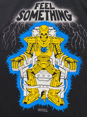 A close up of the artwork on a black short sleeve tee that reads Feel something at the top with silver color and lighting around it. Below the words is a cartoon skull robot in gold that is strapped in an electric chair. Between it's feet is a gold mouse coming out of the bottom of the chair. Crocodile Jackson signature at the bottom.