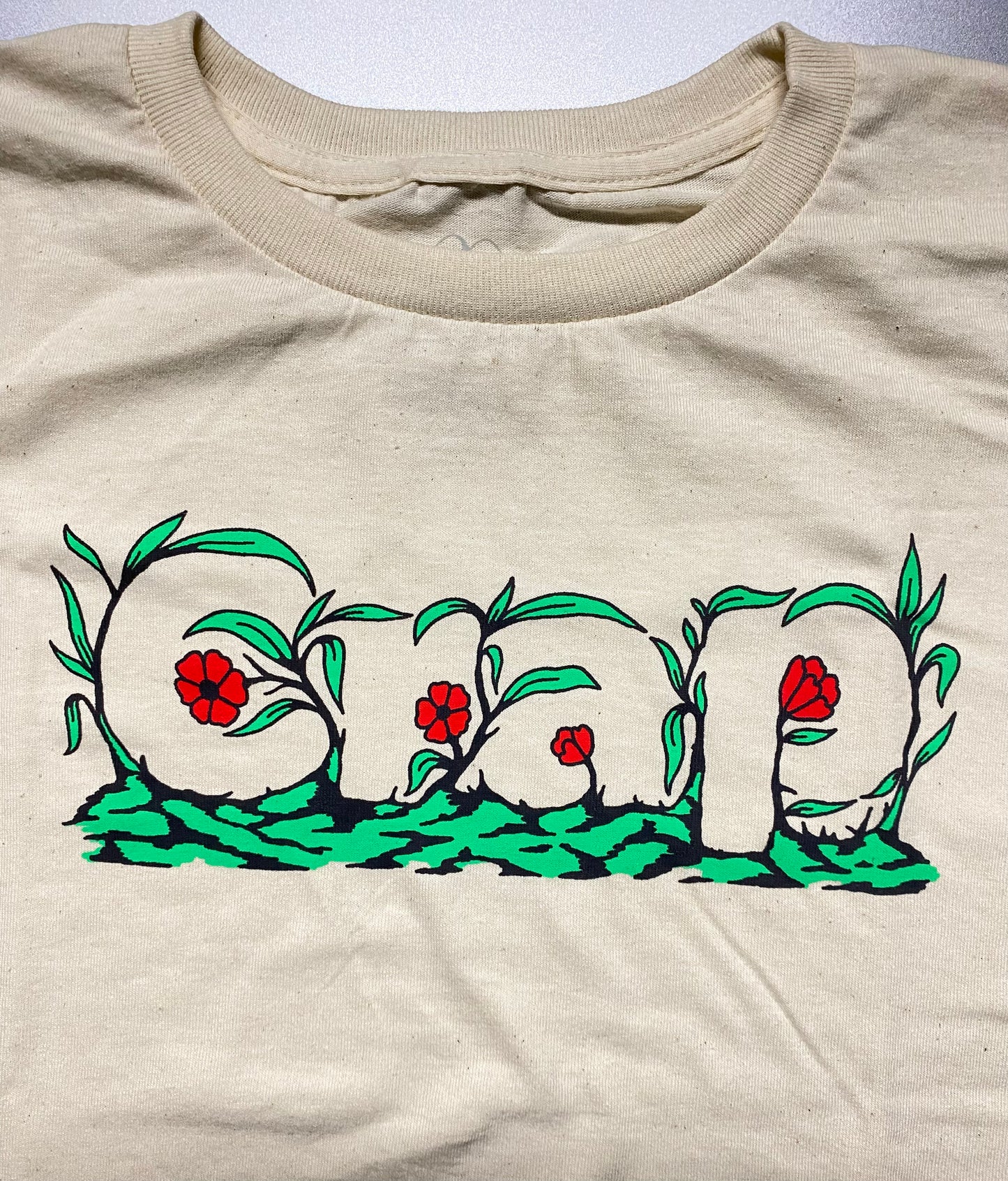 A close up of a cream colored short sleeve tee with the word "Crap" printed on the chest area that is camouflaged in green leaves and red flowers.