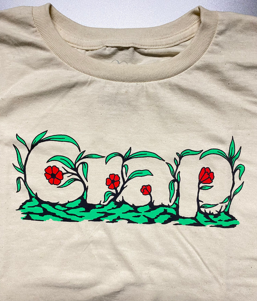 A close up of a cream colored short sleeve tee with the word "Crap" printed on the chest area that is camouflaged in green leaves and red flowers.