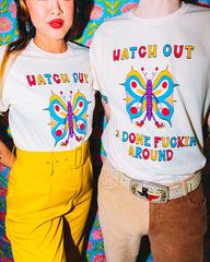 Models in tees.  A cartoon butterfly is in the center and it is wearing white gloves and red tennis shoes. It's wings are gold, blue and pink. Above the butterfly it reads Watch Out in alternating colors of pink, gold & blue. At the bottom it reads I'm Done Fuckin' Around also in the same alternating colors.