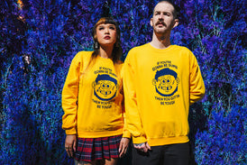 Two models in yellow sweatshirts with a navy print of a cartoon face of a boy. The boy has a swollen right eye with 2 tears. His left cheek has stiches and he's missing a front tooth. His left ear has a piece missing. His dark hair is a mullet cut. The top of the print reads If You're Gonna Be Dumb and the bottom reads Then You Gotta Be Tough.  The backdrop is giant blue shrub.