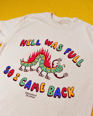 A cream colored t-shirt that reads Hell Was Full at the top and So I Came Back at the bottom in alternating red, yellow, green and blue colored letters. The center is a a smiling cartoon inch worm with 6 legs and tennis shoes for its feet. The top of the worm has flames shooting up and it has two arms in the air showing off its muscles.