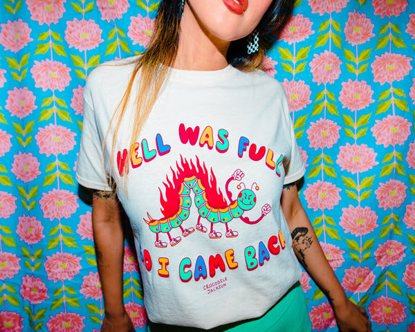 Model wearing a  cream colored short sleeve t-shirt that reads Hell Was Full at the top and So I Came Back at the bottom in alternating red, yellow, green and blue colored letters. The center is a a smiling cartoon inch worm with 6 legs and tennis shoes for its feet. The top of the worm has flames shooting up and it has two arms in the air showing off its muscles.