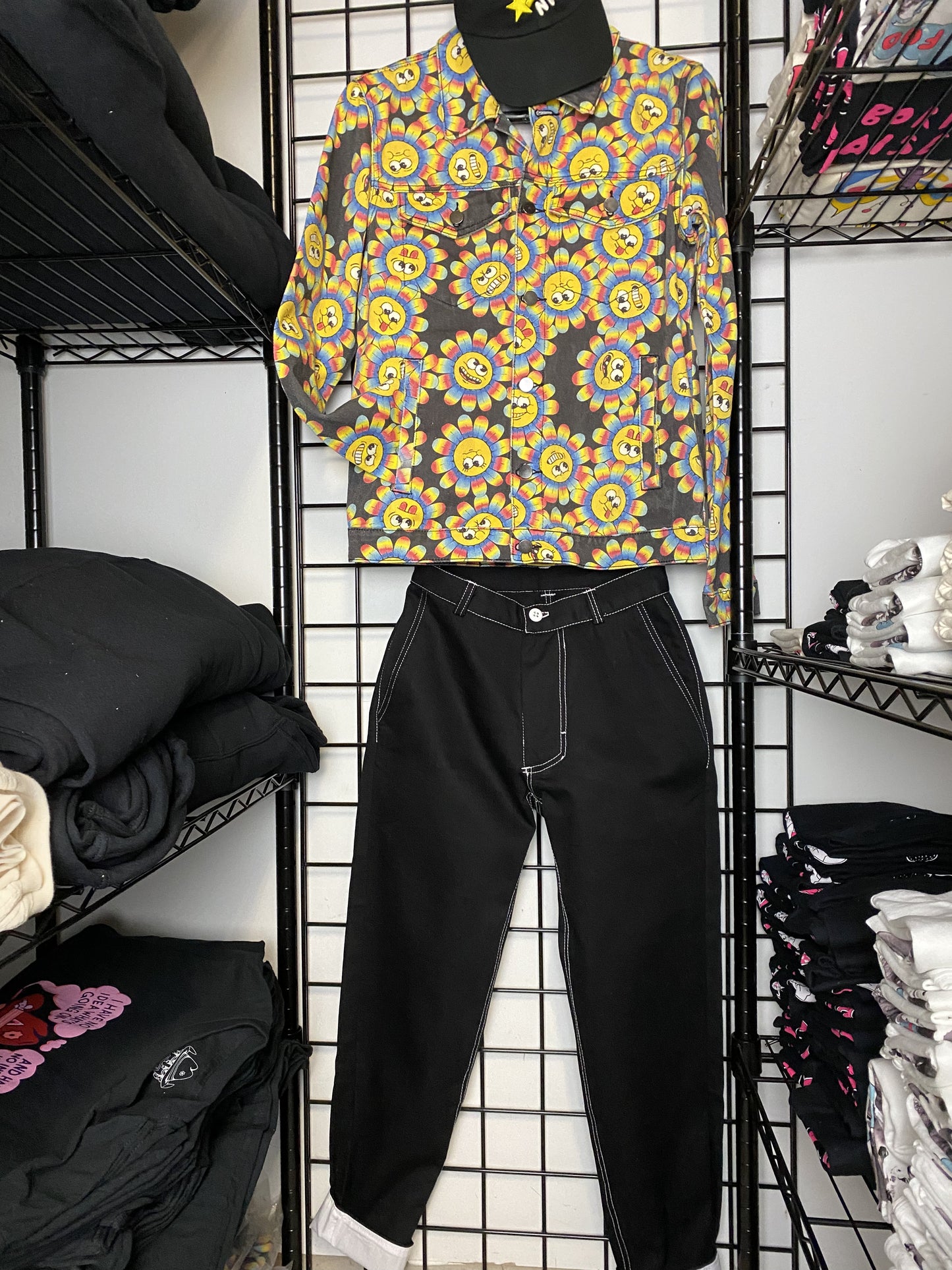 A picture of a black pair of pants (front view) hung on a black wire mesh background.  The top stitching is white as well as the button.  The cuffs are rolled up to show the white fabric underneath.  Above the paints is a jacket with a print of flowers with smiley faces.  Above the jacket is a back baseball cap.