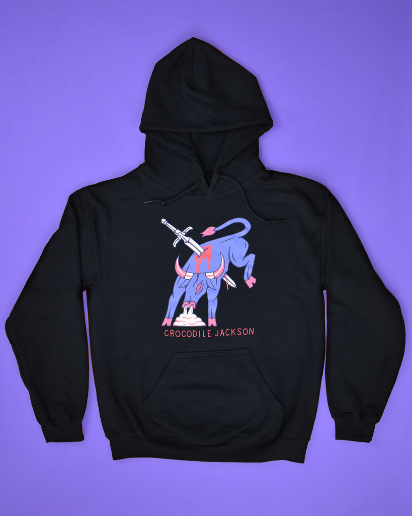 Flat-lay image of a black hoodie on a periwinkle background. The hoodie features art of a periwinkle colored bull in full fight motion, bucking it's back legs with steam coming out of his nose. There is a sword piercing the body of the bull and it is bleeding. At the bottom of this art, it reads Crocodile Jackson.