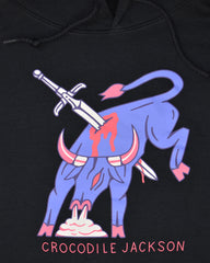Close-up image of a black hoodie. The hoodie features art of a periwinkle colored bull in full fight motion, bucking it's back legs with steam coming out of his nose. There is a sword piercing the body of the bull and it is bleeding. At the bottom of this art, it reads Crocodile Jackson.