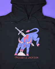 Close-up image of a flat laying black hoodie with a periwinkle background. The hoodie features art of a periwinkle colored bull in full fight motion, bucking it's back legs with steam coming out of his nose. There is a sword piercing the body of the bull and it is bleeding. At the bottom of this art, it reads Crocodile Jackson.