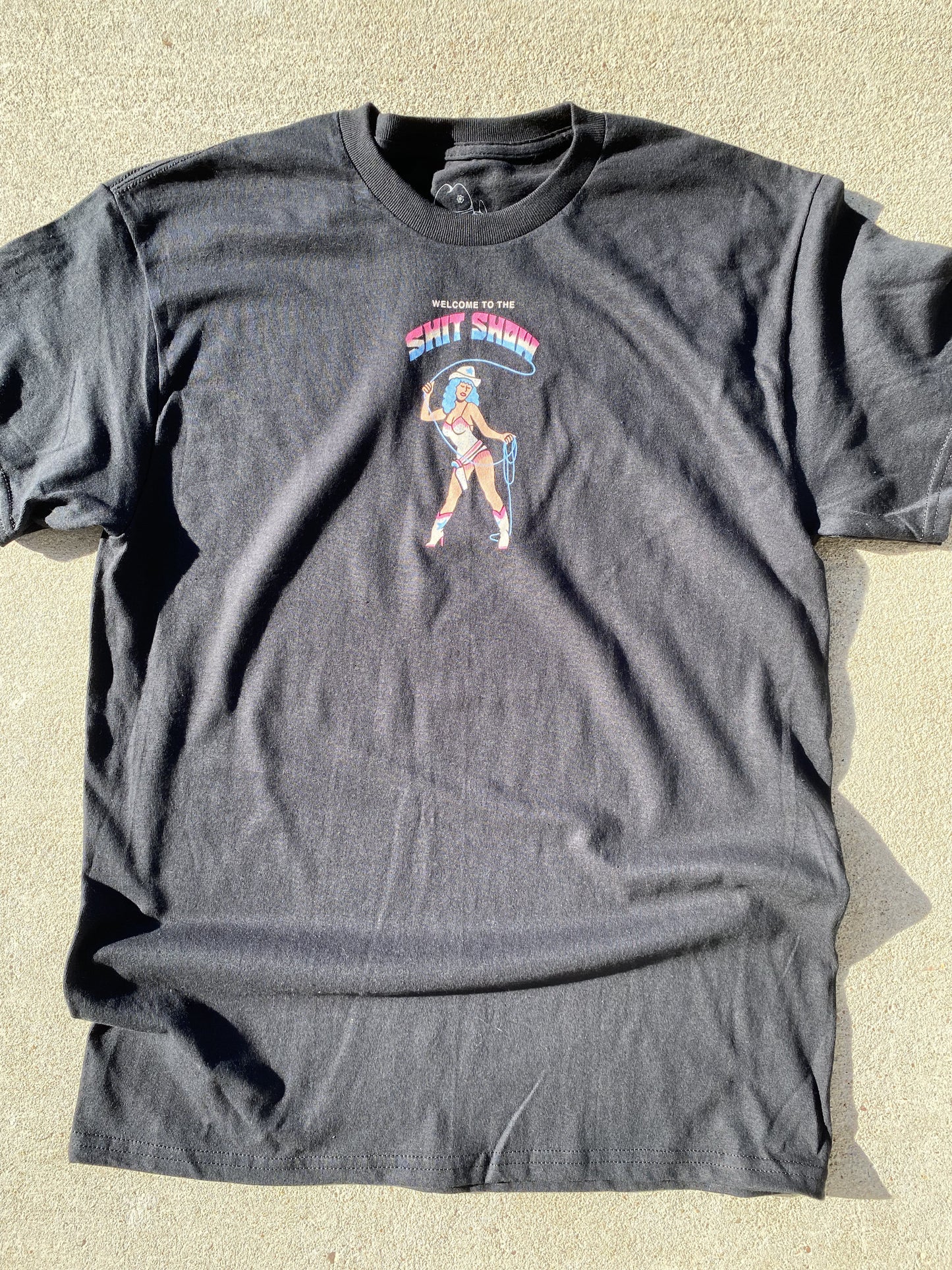 A black short sleeve t-shirt that reads Welcome to the Shit Show. The last 2 words are in a large font in rainbow colors of light blue, white and pink. Below the words is a cowgirl with blue hair in a white bathing suit and white boots. She is holding a blue lasso and have a holster with a pistol.  Background is concrete.