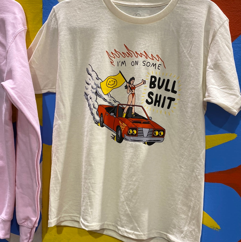A short sleeve natural colored t-shirt with a chest print of a cartoon girl in a red bikini standing on a red convertible that is driving itself. The car has gold flames above the front tires and smoke coming off the back wheels. The girl is holding a yellow flag with a smiley face on it. Above her reads I'm On Some Bull Shit with red outline flames on top.