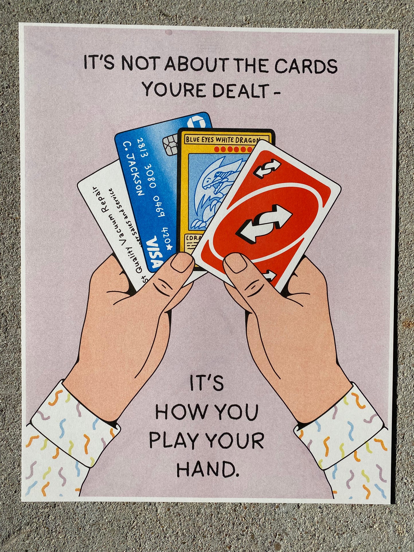 A poster with a light purple background and 2 cartoon hands holding 4 cards - a business card, a credit card and 2 game cards.  The sleeves are white with a multi-colored design that looks like small worms.  The top of the poster reads "It's Not About the Cards You're Dealt - It's How you Play Your Hand".