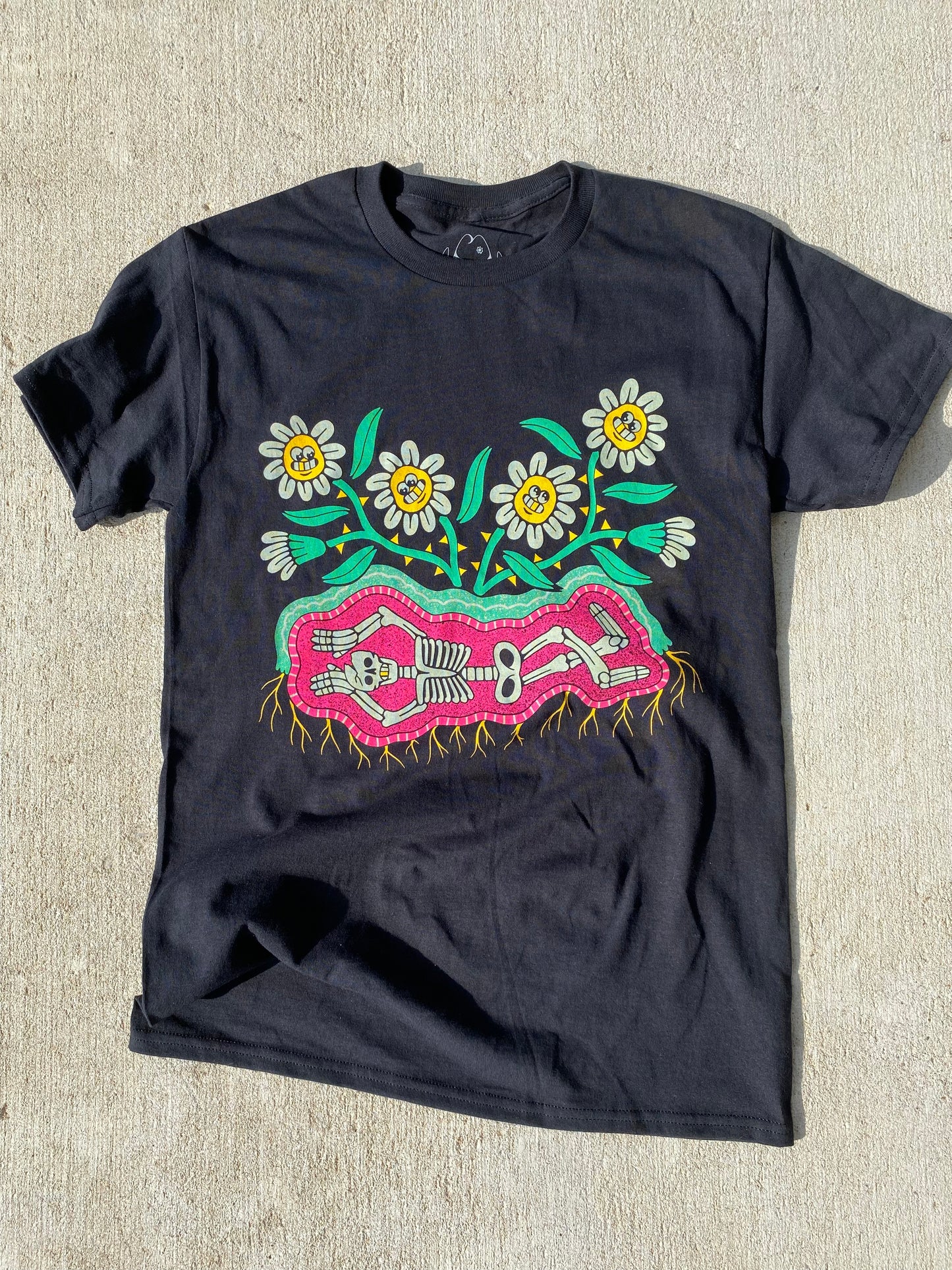 Black short sleeve t-shirt with 6 cartoon flowers with white petals and smiling faces. Below the flowers is a skeleton laying down in pink dirt and roots growing out of the bottom.