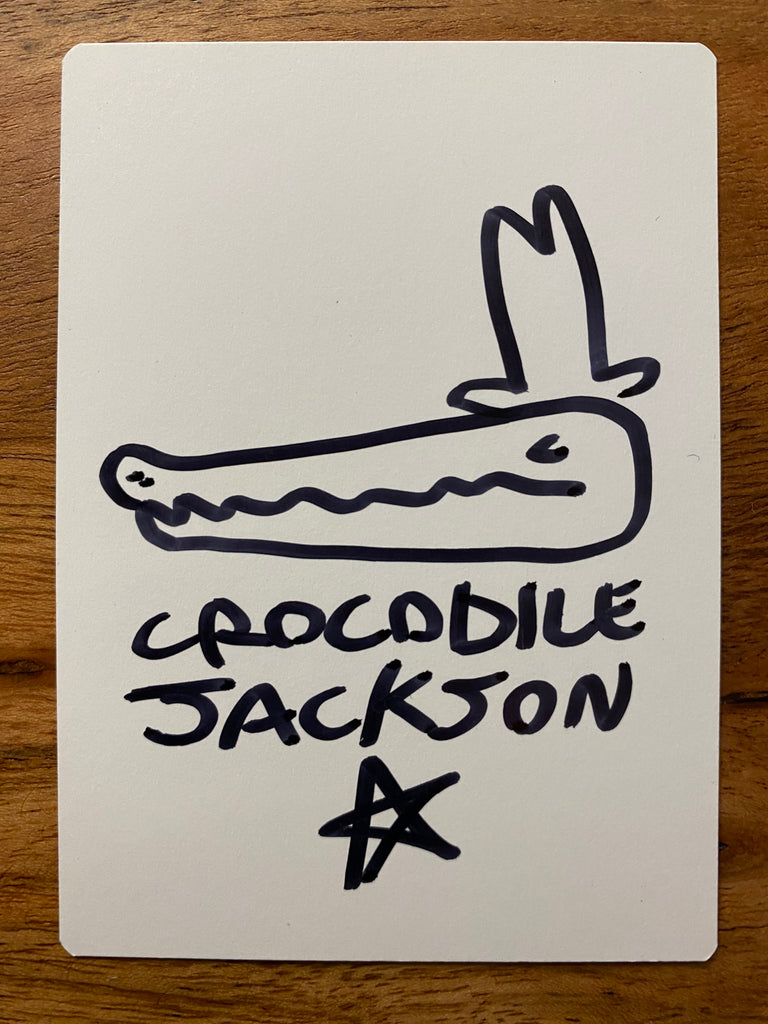 The back of the trading card that is all white where the artist has hand drawn a crocodile head with a tall cowboy hat and below that is the artist signature - Crocodile Jackson.