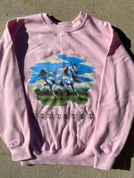 Pink sweatshirt with a horse printed in the middle & it is walking on green grass under a blue sky with white clouds. Above the horse are the words Life is Weird and below the horse it reads And That Is Ok. Below those words are 8 skulls lined up.  Background is concrete.