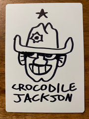 The back of the card that is all white where the artist hand drew a smiling cowboy head with his signature below it - Crocodile Jackson - in black marker.