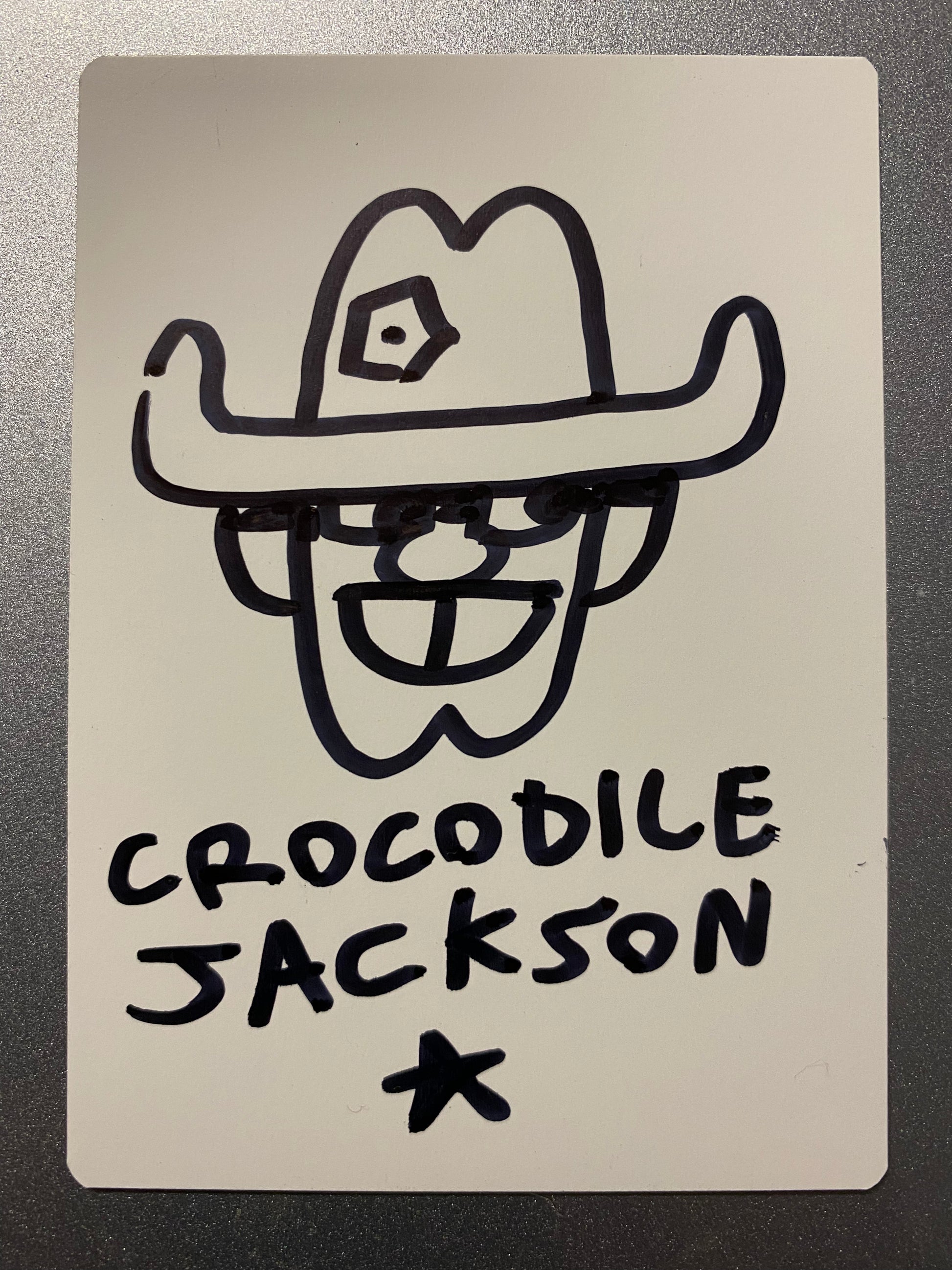 The back of the trading card that is all white where the artist has hand drawn a smiling cartoon cowboy head.  There is a bullet hole in his hat.  At the bottom is the artist signature - Crocodile Jackson - with a star below it.  All in black marker.
