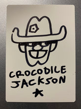Load image into Gallery viewer, The back of the trading card that is all white where the artist has hand drawn a smiling cartoon cowboy head.  There is a bullet hole in his hat.  At the bottom is the artist signature - Crocodile Jackson - with a star below it.  All in black marker.
