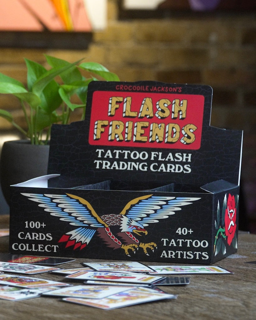 A black box of trading cards that reads Flash Friends Tatttoo Flash Trading Cards.  The front side of the box has a cartoon eagle with wings spread.  The box is on a wood table with some of the single cards spread out.  Behind the box is a plant in a grey pot.